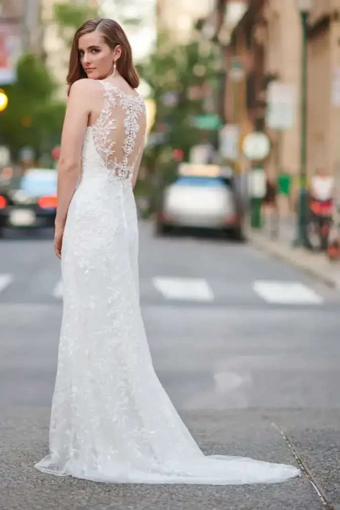 A woman in a lace wedding dress with an intricate back design stands confidently on a city street. The scene conveys elegance and urban charm.