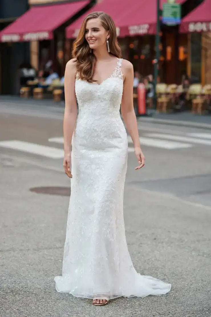 A woman in a white lace wedding dress walks confidently on a city street, smiling. Red awnings and outdoor cafe tables are in the background.