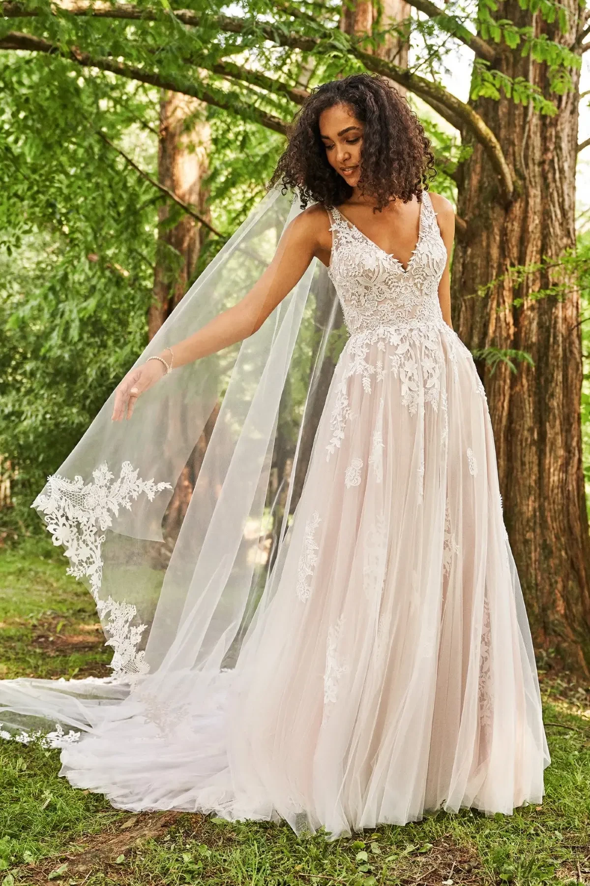 A bride in a lace wedding gown and veil stands in a sunlit forest. The dress has intricate lace details, and her serene expression conveys joy and elegance.