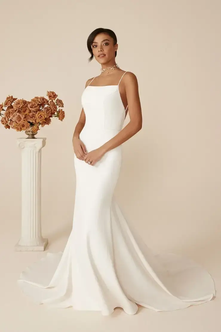A model wearing a sleek, white wedding dress with a fitted silhouette standing next to a vase of dried flowers on a pedestal.