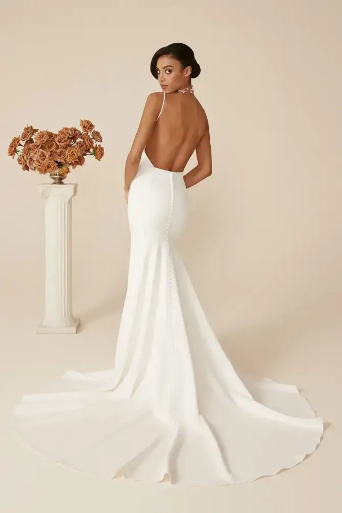 A woman in a backless white gown poses with her back to the camera, showcasing a flowing train, with a decorative vase of dried flowers in the foreground against a neutral background.