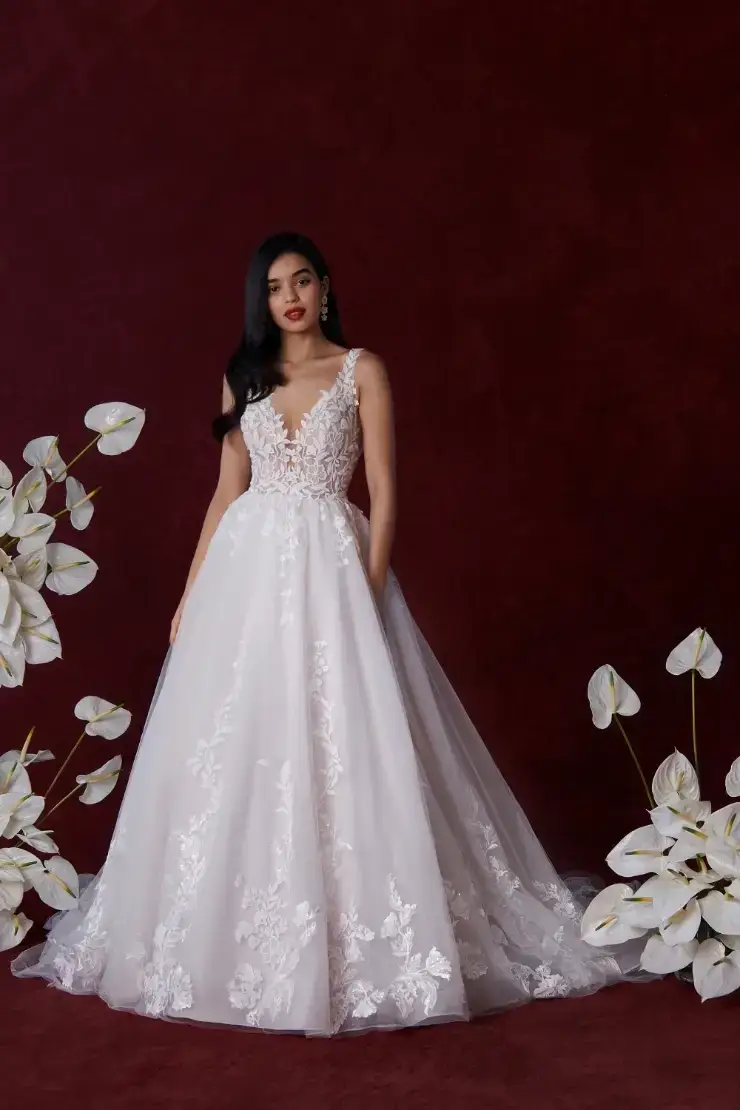 A woman in a white gown with floral details stands against a deep red background, surrounded by white flowers.