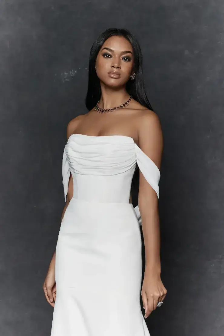 A person wearing a strapless white dress with draped sleeves, standing confidently against a dark background.