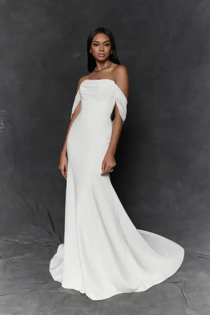 A model wearing a white off-shoulder gown with a flowing train, standing against a dark background.