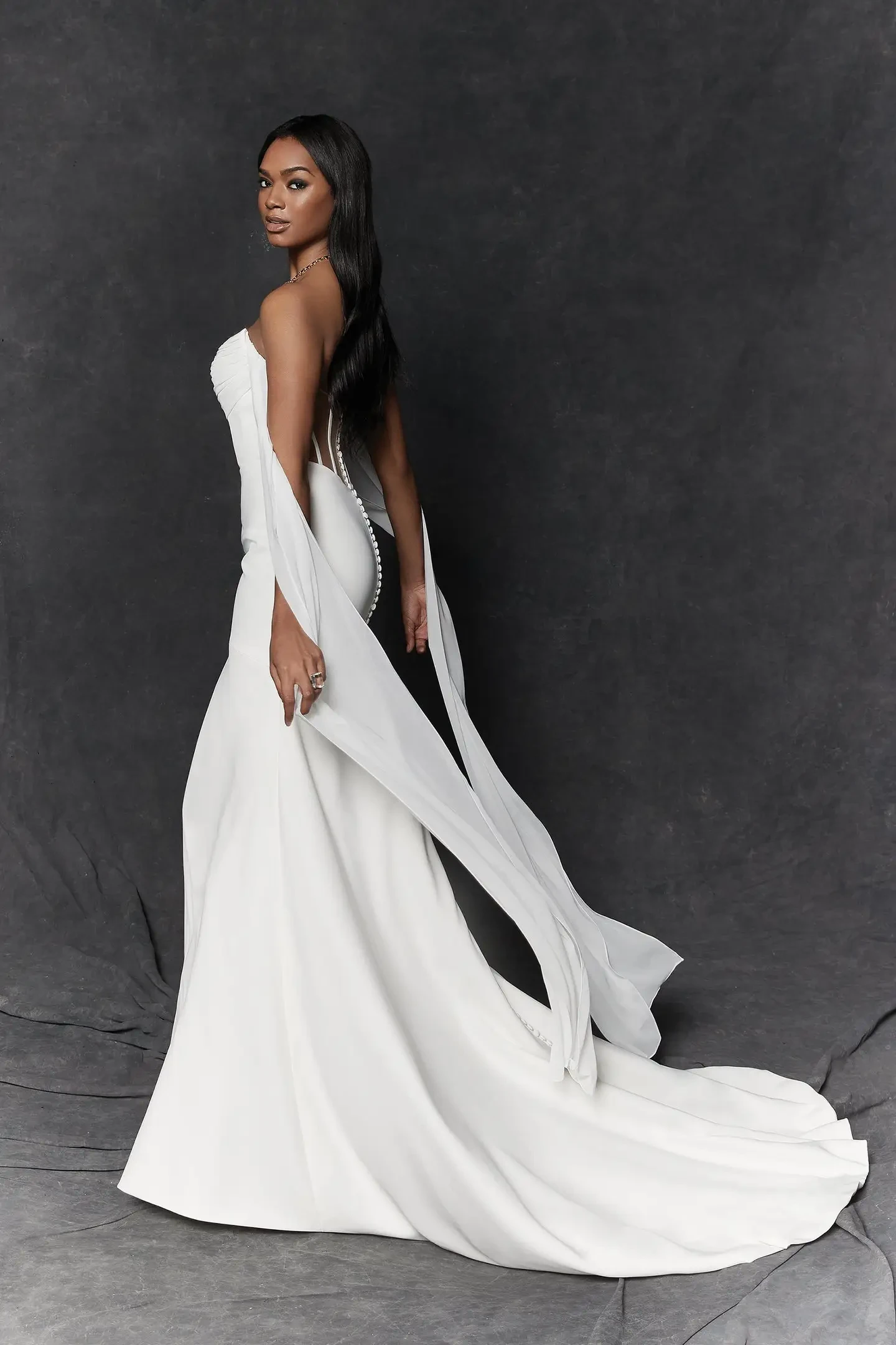 Model is wearing a modern crepe fit and flare gown featuring a ruched chiffon neckline, detachable streamers tied into a bow over an illusion back with exposed boning and a chapel-length train.