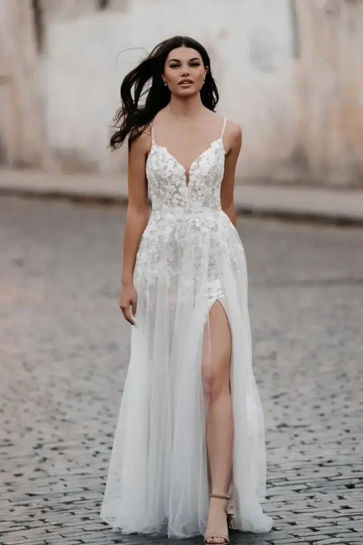 A woman confidently walks on a cobblestone street, wearing a flowing white gown with floral details and a thigh-high slit. The background is a blurred wall.