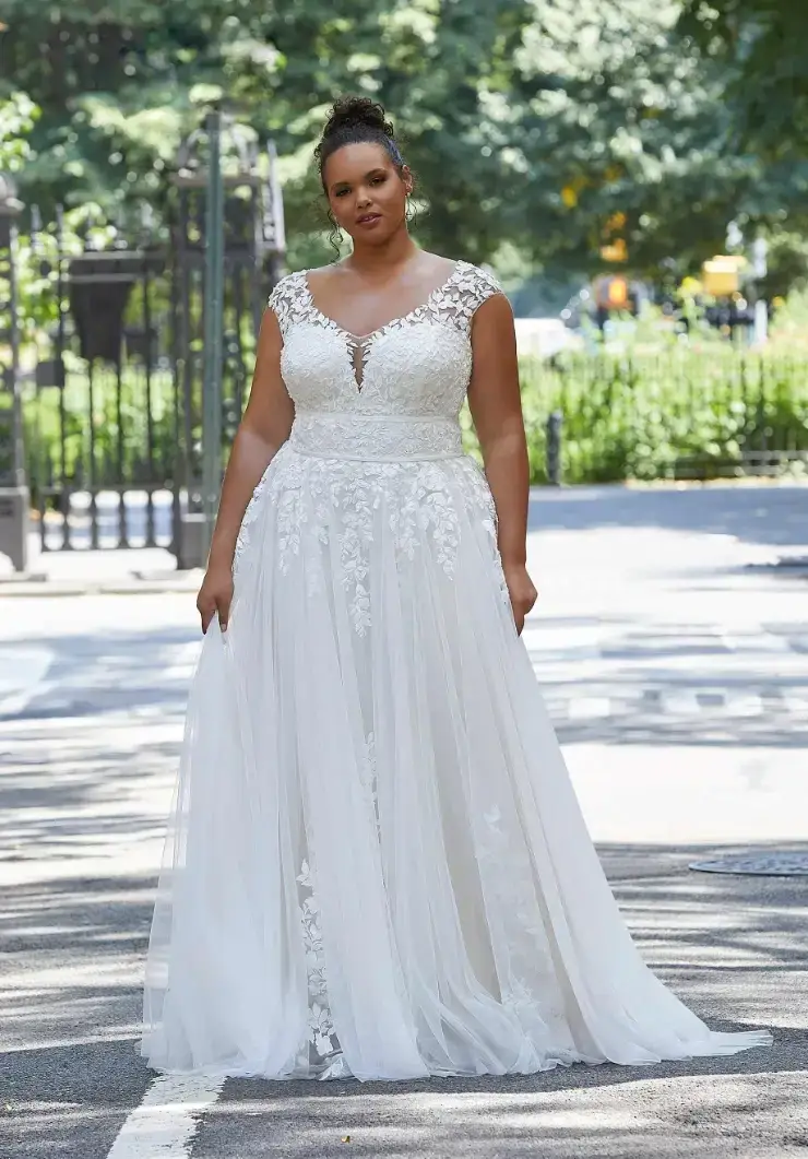 A woman in a lace wedding dress stands confidently on a sunlit path, surrounded by lush greenery and a serene park backdrop.