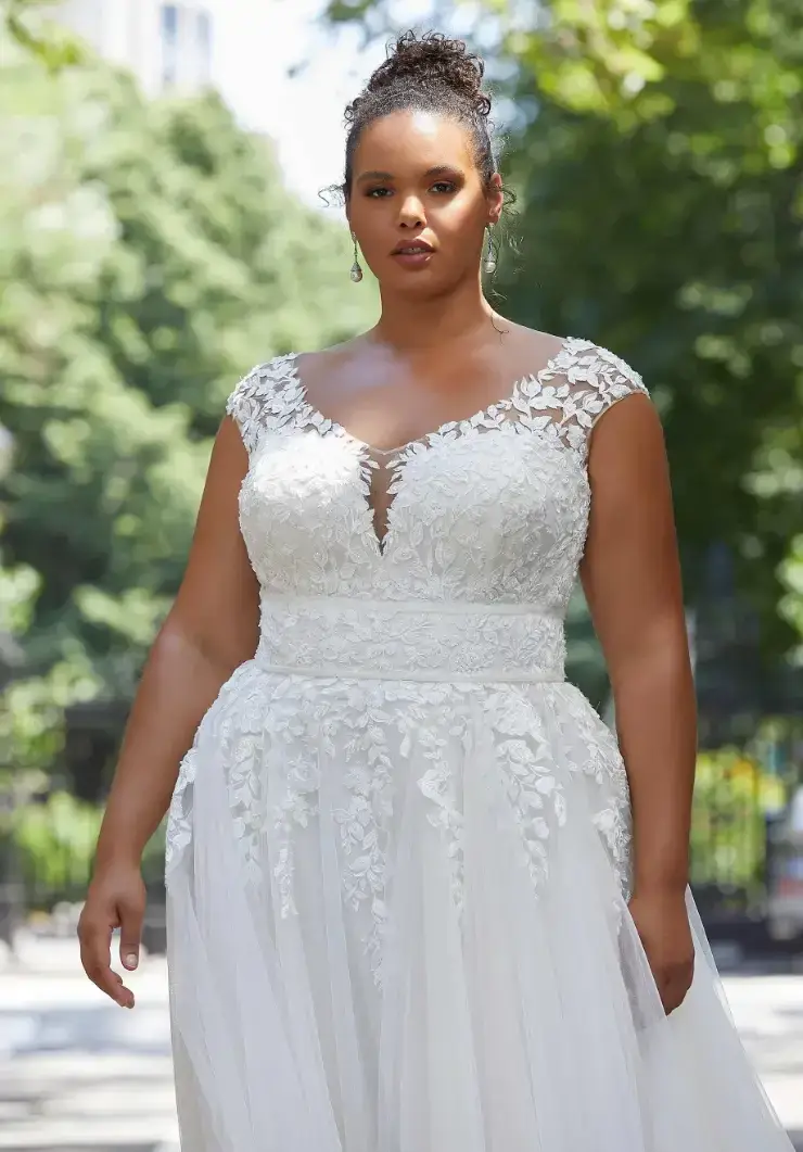 A woman stands outdoors wearing a white lace wedding dress with floral patterns, exuding elegance and confidence. Lush green trees are visible in the background.