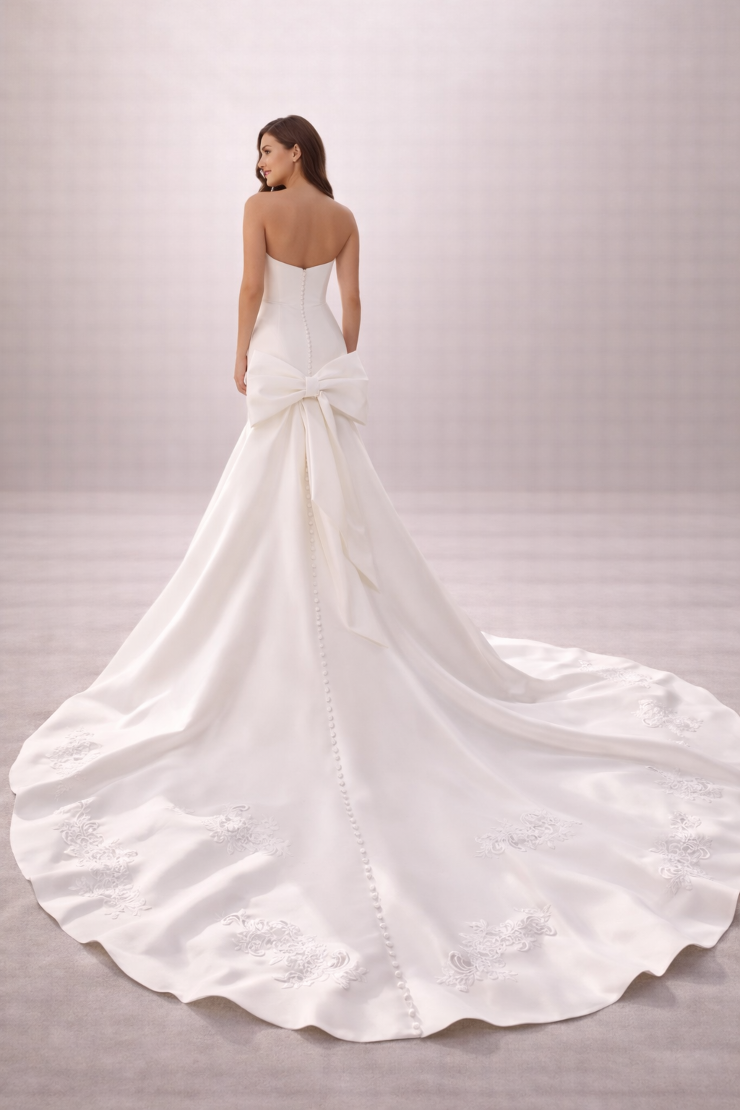 A woman in a white wedding dress with a long train is seen from the back, featuring a strapless design and large bow at the waist.
