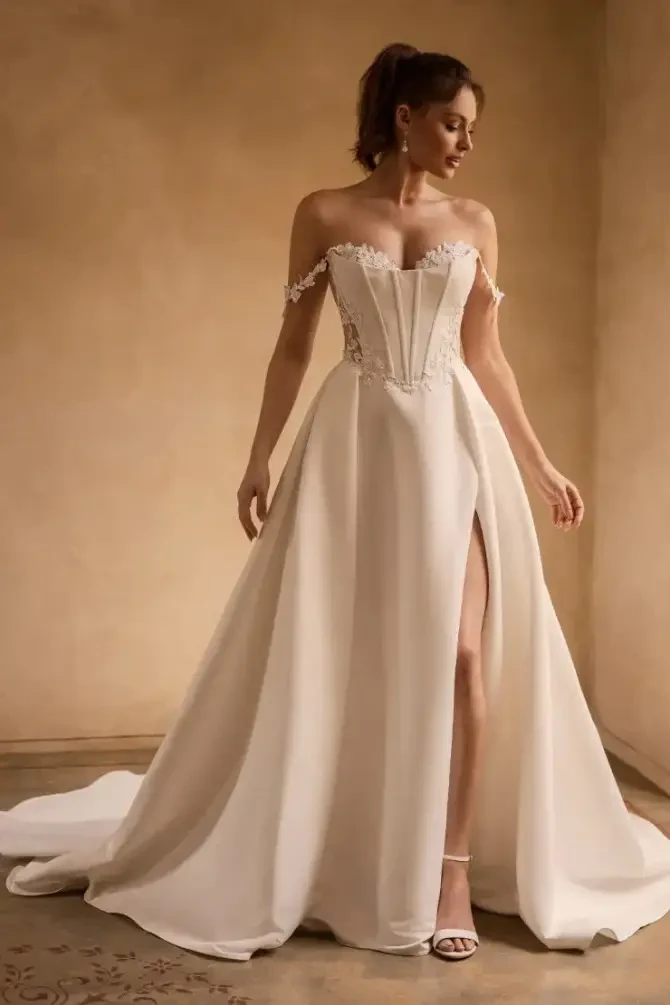 Model is wearing a dove satin ballgown with an illusion boned bodice, off-shoulder lace straps, a leg slit, and a dramatic illusion back with fabric-covered buttons.