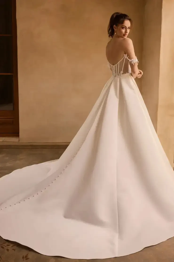 Model is wearing a dove satin ballgown with an illusion boned bodice, off-shoulder lace straps, a leg slit, and a dramatic illusion back with fabric-covered buttons.