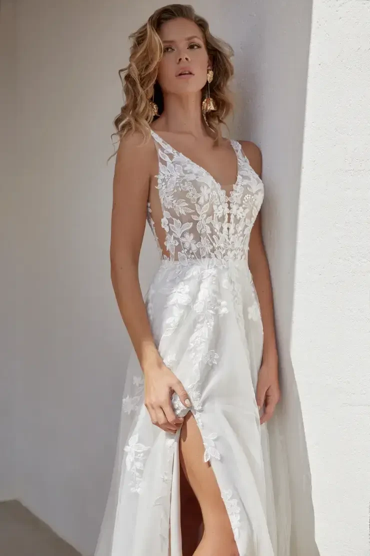 A woman in a romantic white gown with a high slit, standing against a white wall. The dress features delicate floral details and a fitted bodice, complementing her elegant pose.