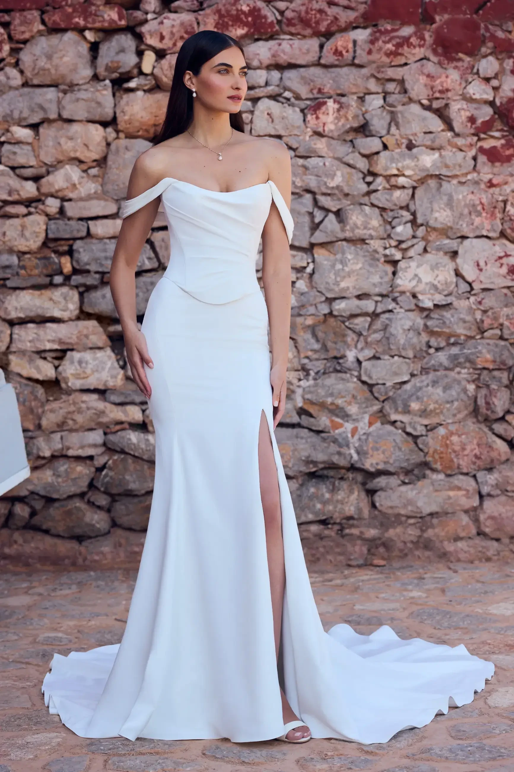 Model is wearing a minimalist matte satin sheath gown with a scoop neckline, off-shoulder straps, a thigh-high slit, and a romantic corset lace-up back.
